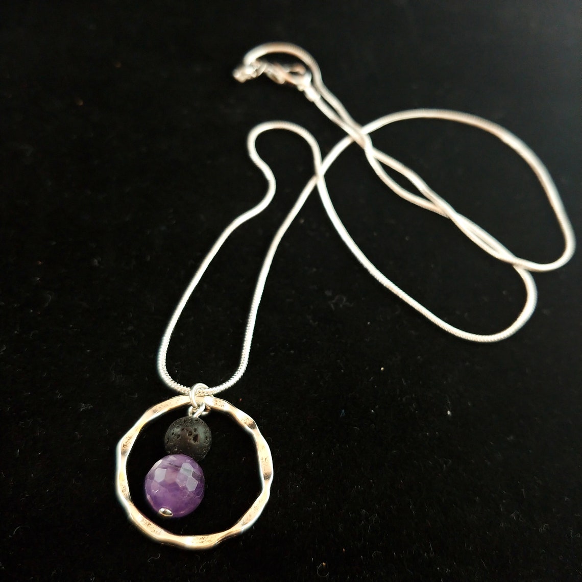 Silfur hálsmen með amethyst og lava steinum. Steinarnir hanga fyrir miðju í grófum silfur hring. Hálsmenið liggur á svörtum bakgrunn.